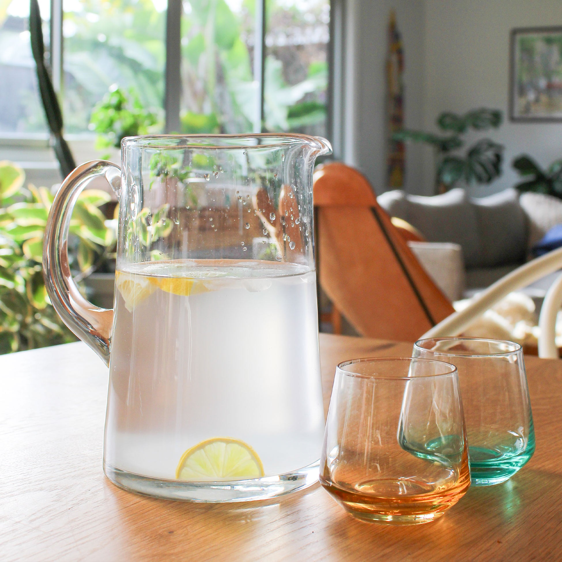 Clear glass pitcher with lemon slices on a wooden table, surrounded by two glasses.