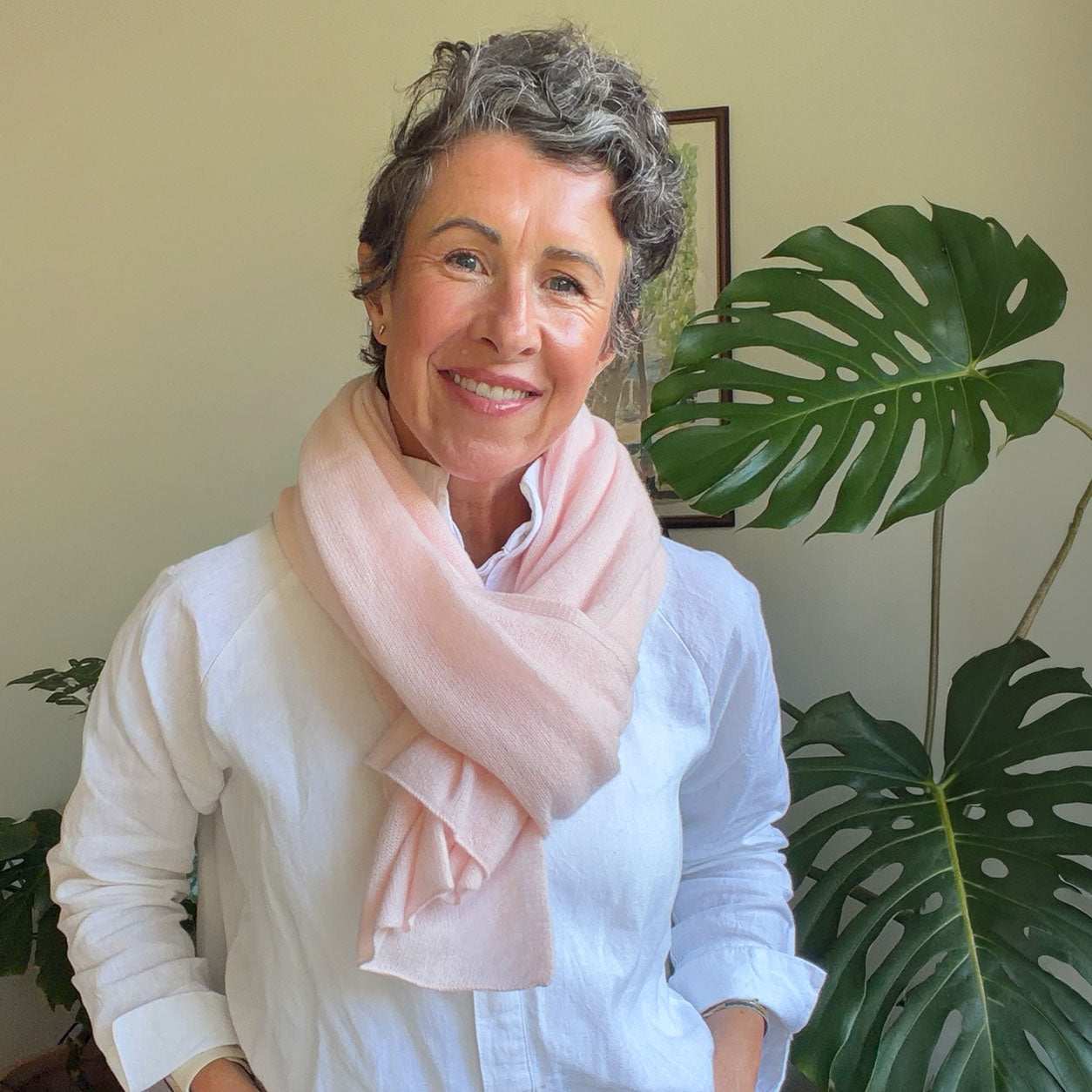 A smiling woman with short gray hair wearing a soft light pink scarf and a white button-up shirt, standing indoors next to a large green monstera plant.