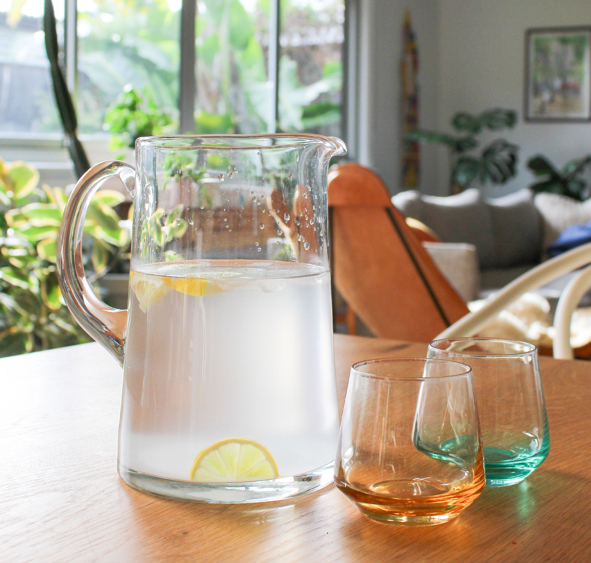 Clear glass pitcher with lemon slices on a wooden table, surrounded by two glasses.