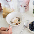 Amy adding maca powder to a bowl of muesli with blueberries on a countertop.