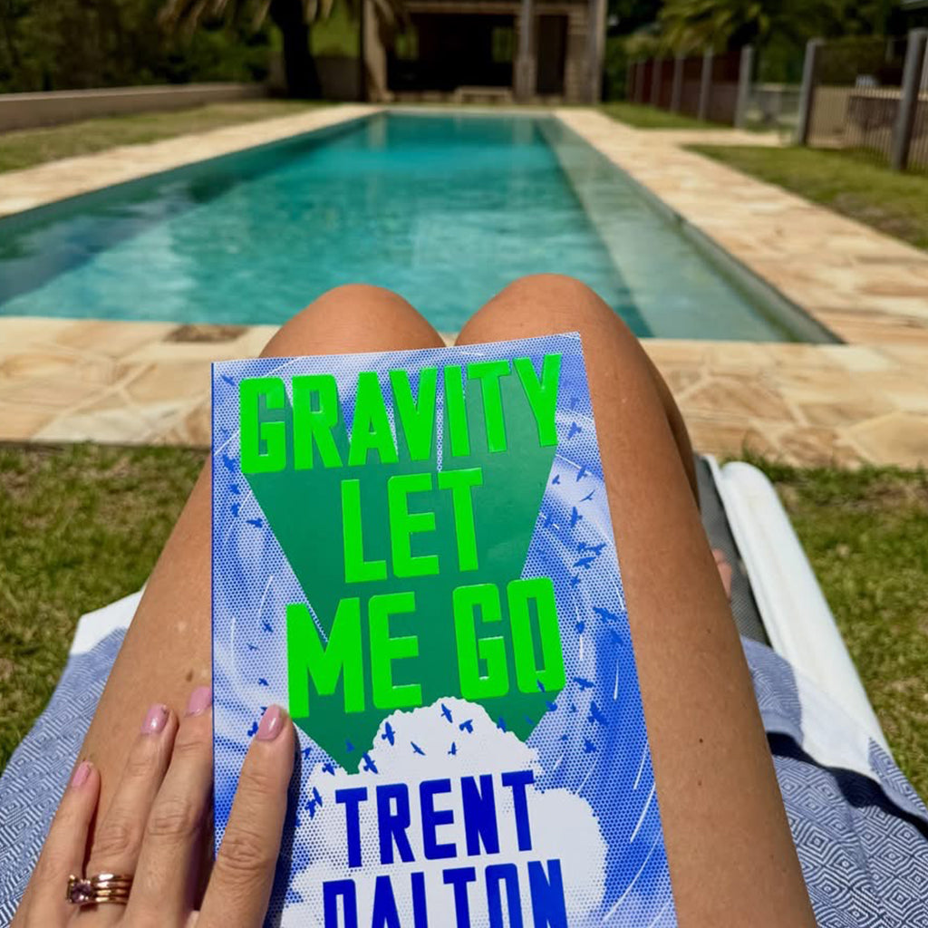 Amy holding a book titled 'Gravity Let Me Go' by Trent Dalton by a pool.
