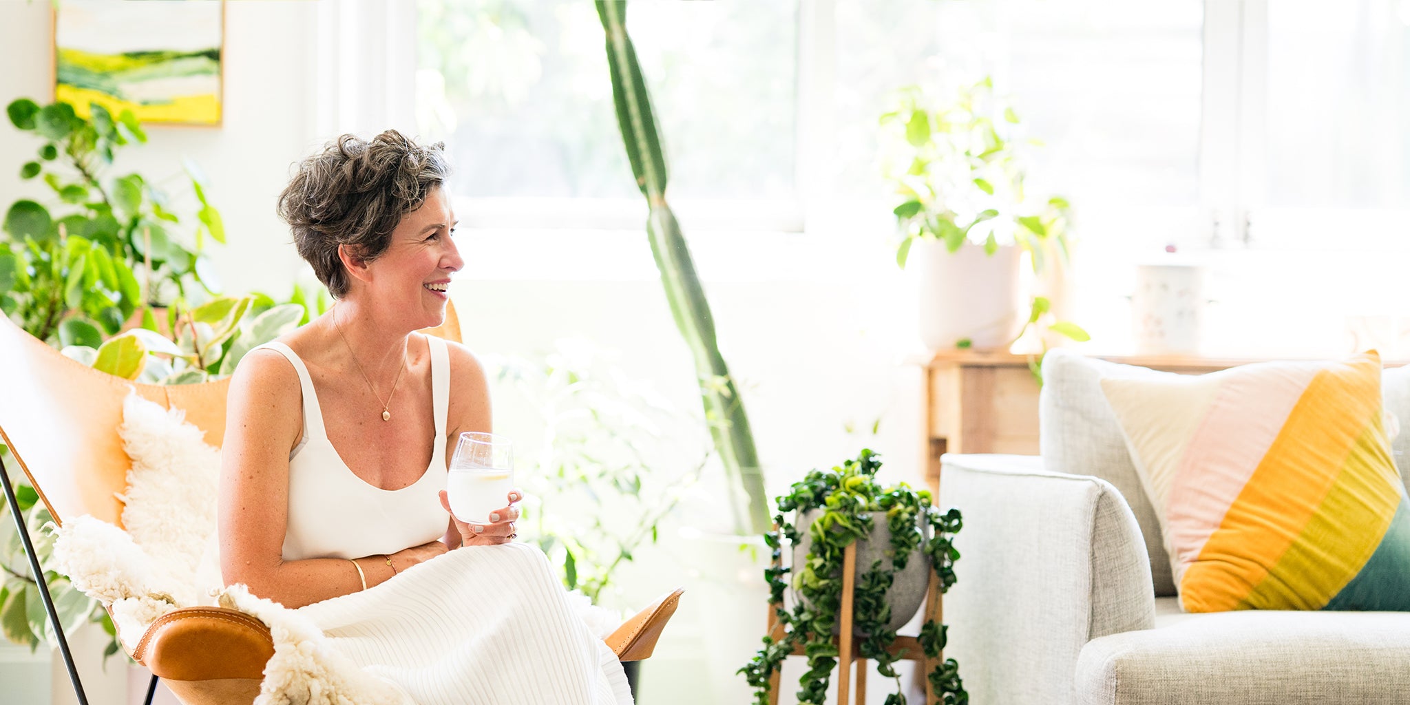 Amy in a white dress sitting on a couch in a bright, plant-filled room.